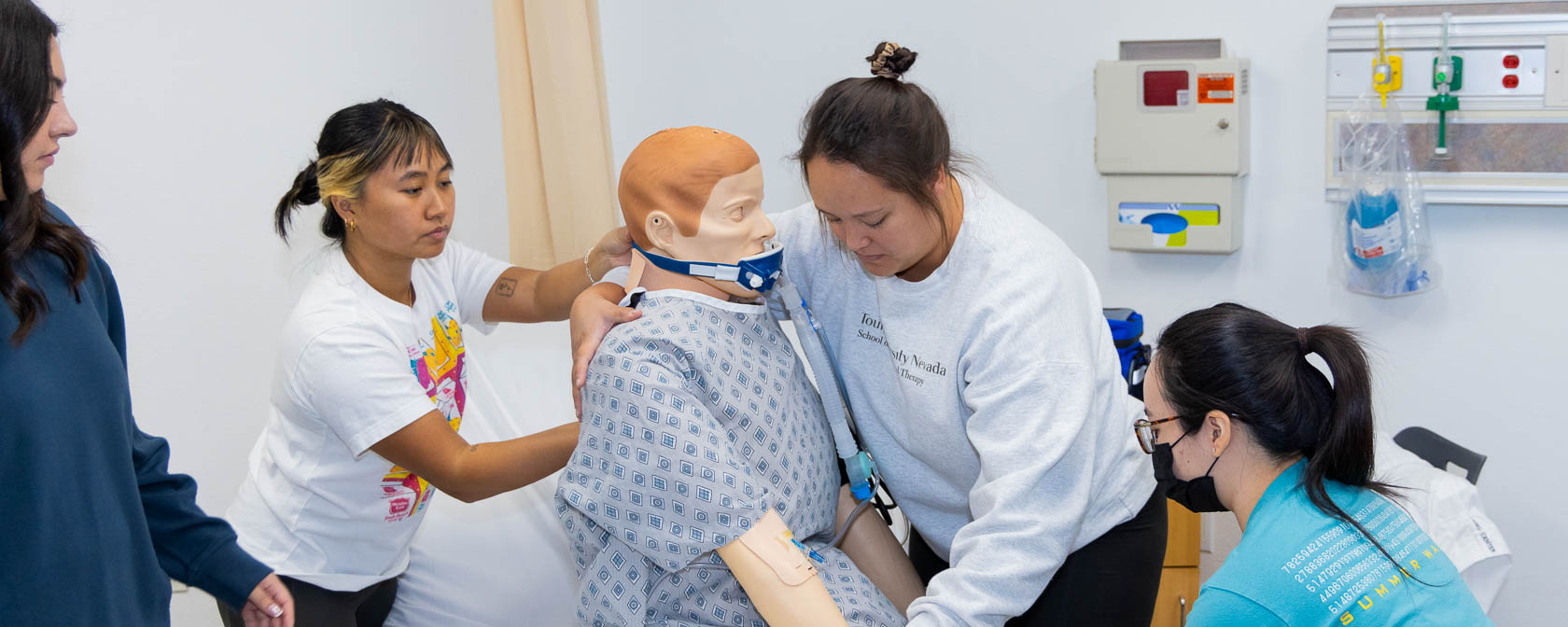 Four students work as a group to adjust a simulation manikin wearing a gown in a hospital bed.