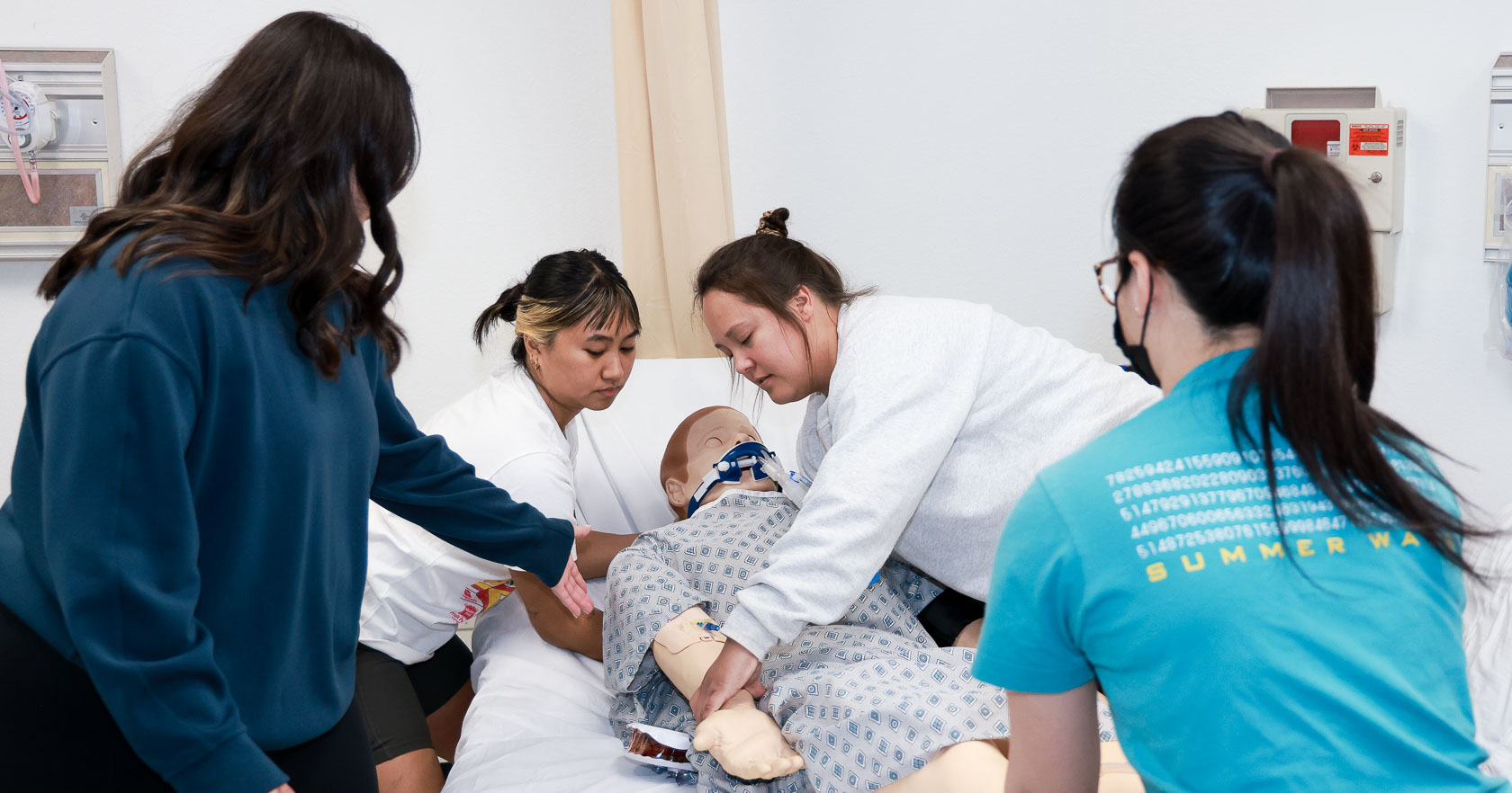Four students move a simulation mannequin in a gown from a hospital bed. One student guides the others, while two student lift the upper body, and one student holds the feet.