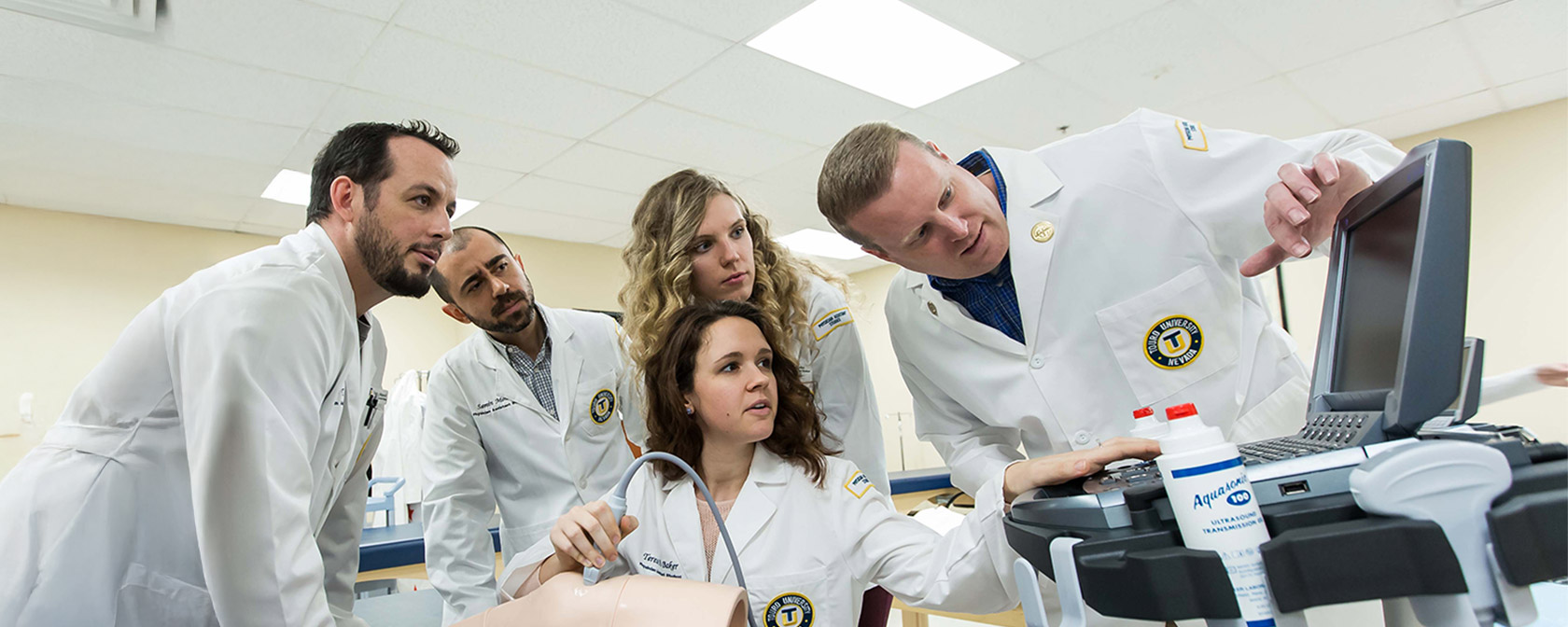 A faculty member in a white coat points to a monitor while a student uses an ultrasound device on a medical trainer's leg. A group of students in white coats observes the monitor.