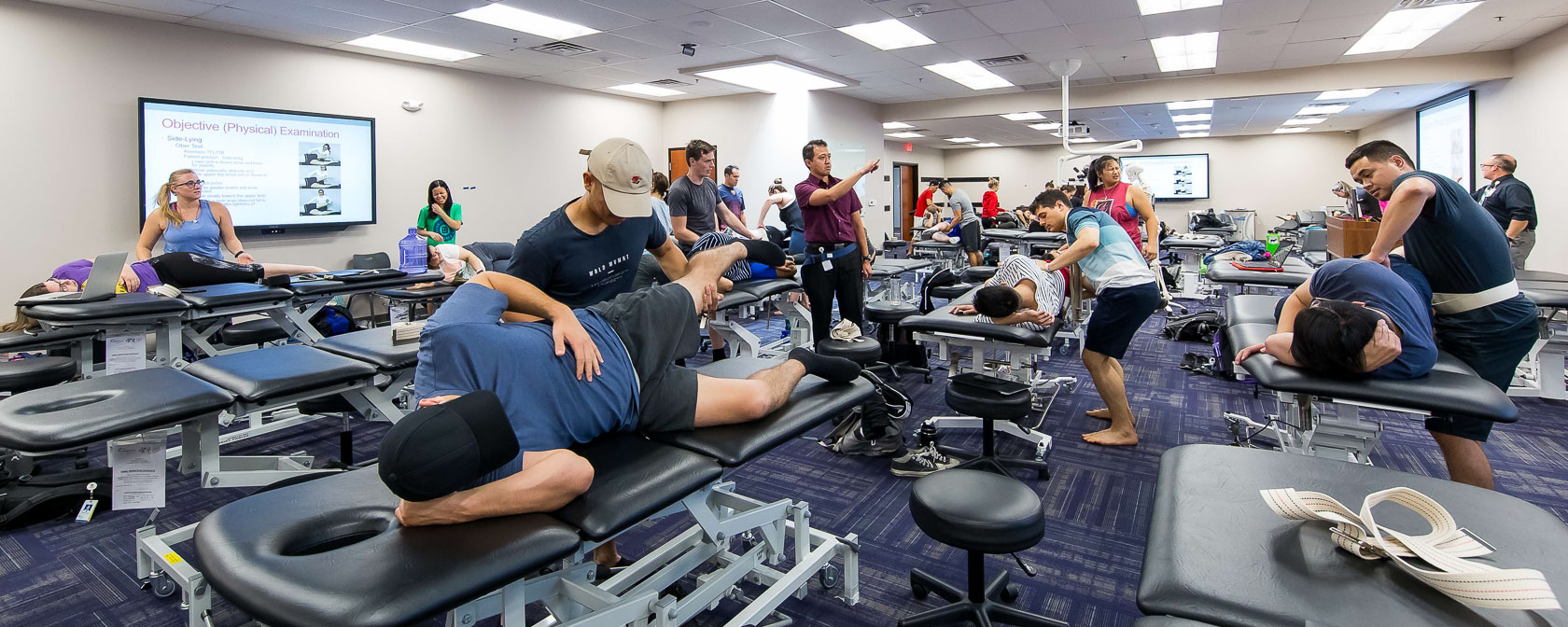 Students in pairs practice physical exams on each other in a classroom. One student lies on the exam table while the other checks their leg and lower back.