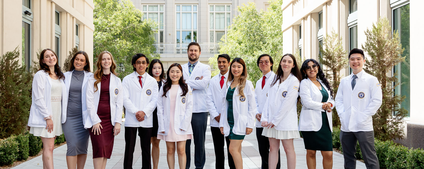 A group of medical students wearing white coats and dress attire pose for a group photos in an outdoor courtyard.